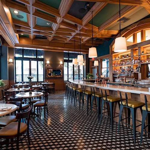 A stylish restaurant interior with patterned flooring, wooden ceiling, bar counter, stools, and tables set for dining, warmly lit ambiance.