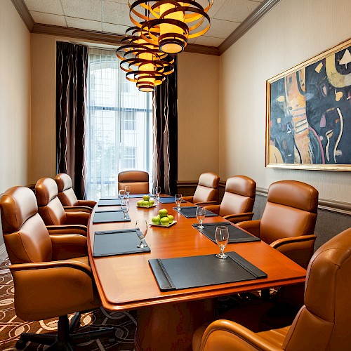 A conference room with a wooden table, leather chairs, a painting on the wall, and overhead lights, featuring notepads and glasses on the table.