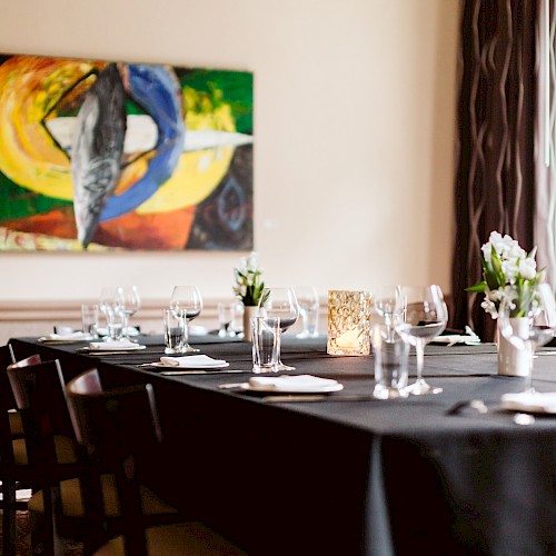 A dining table with a black cloth, set for a meal, features glasses and napkins. A colorful abstract painting hangs on the wall.