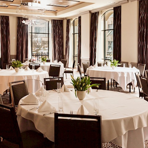 The image shows a sophisticated dining area with round tables, white tablecloths, green plants, elegant chairs, and large windows.