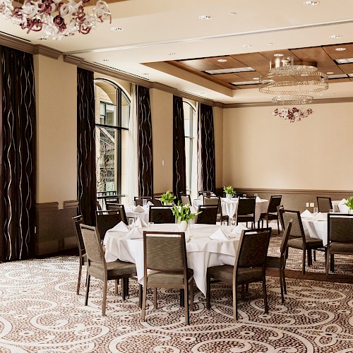 The image shows a banquet room with round tables, white tablecloths, chairs, and decorative plants, featuring large windows and elegant lighting.