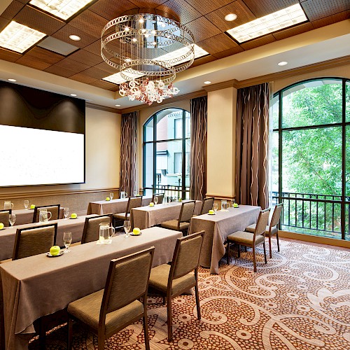 A conference room with tables and chairs facing a blank screen, decorated with elegant lighting and large windows showing greenery outside.