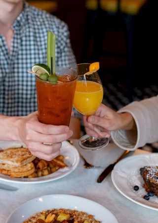 Two people clinking a Bloody Mary and a mimosa over a table with brunch dishes including toast, potatoes, and fruit-topped French toast.