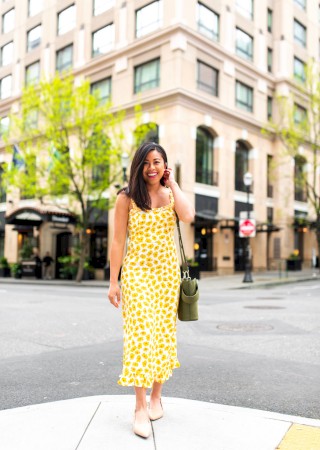 A person in a yellow floral dress stands on a city street, smiling, with a building in the background.