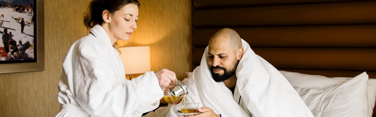 A person in a bathrobe pours tea or similar for another in bed, both in a cozy hotel or home setting, with warm lighting and light wood tones.