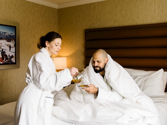 A person pours tea for someone in bed. Both are in bathrobes, suggesting a relaxing morning in a hotel or home setting.