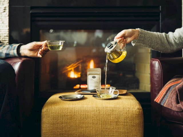 Two people are enjoying tea in front of a fireplace. One pours tea from a teapot, while the other holds a cup.