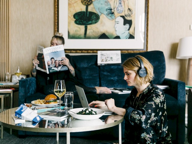 Two people are in a room; one reads a magazine on a couch, the other works on a laptop at a table with food and drinks.
