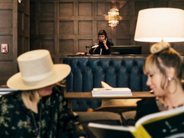 Two people are sitting and reading large books in a cozy room with wood paneling and a front desk in the background.