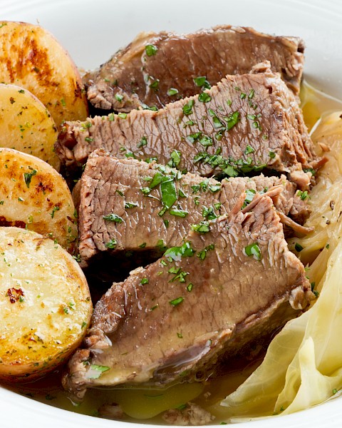 The image shows a dish with slices of cooked beef, roast potatoes, and cabbage garnished with herbs on a white plate.