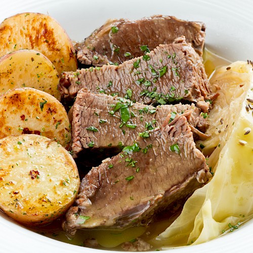 The image shows a dish with slices of cooked beef, roast potatoes, and cabbage garnished with herbs on a white plate.