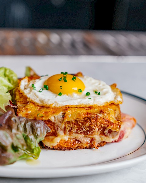 A croque madame sandwich topped with a fried egg, garnished with chives, and served with a side of greens on a plate.