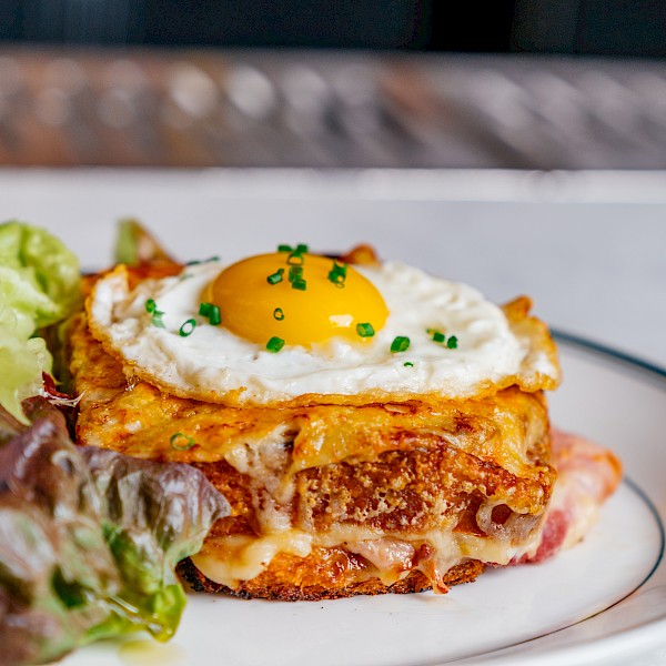 A croque madame sandwich topped with a fried egg, garnished with chives, and served with a side of greens on a plate.