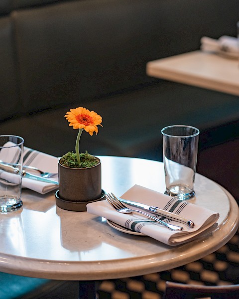 A round table in a restaurant with place settings, glasses, and a potted flower centerpiece, against green seating.