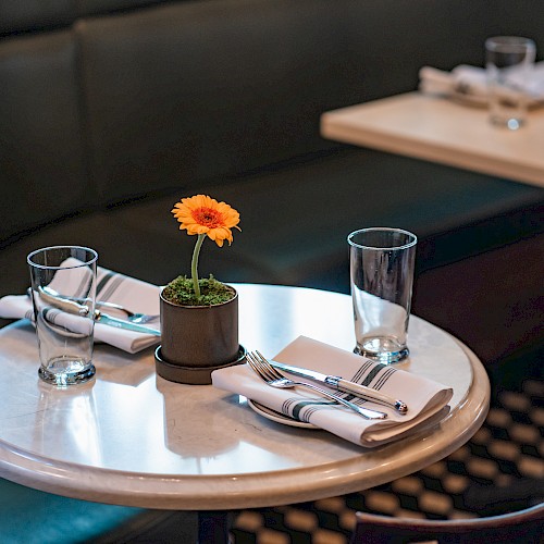 A round table in a restaurant with place settings, glasses, and a potted flower centerpiece, against green seating.