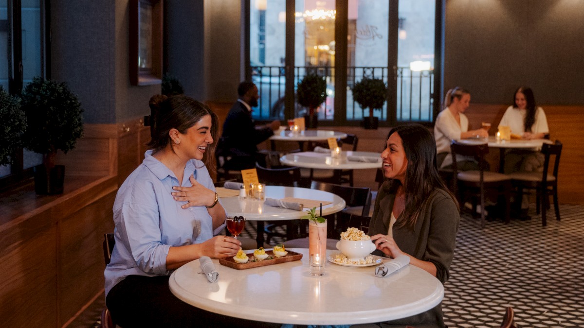 Two women laughing at a restaurant with drinks and snacks on the table, enjoying their time together.