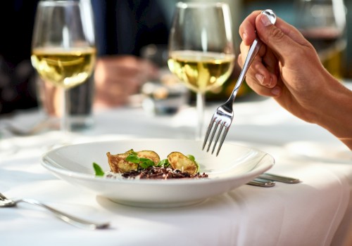 A person holds a fork over a gourmet dish on a white table, accompanied by wine glasses.