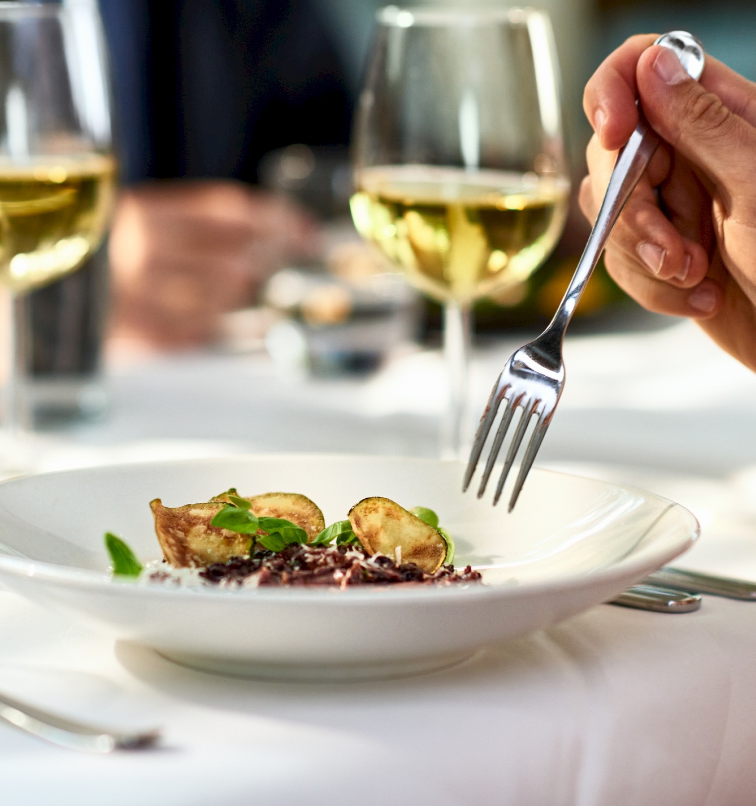 A person holds a fork over a dish with greens and potatoes, with wine glasses on a white tablecloth.