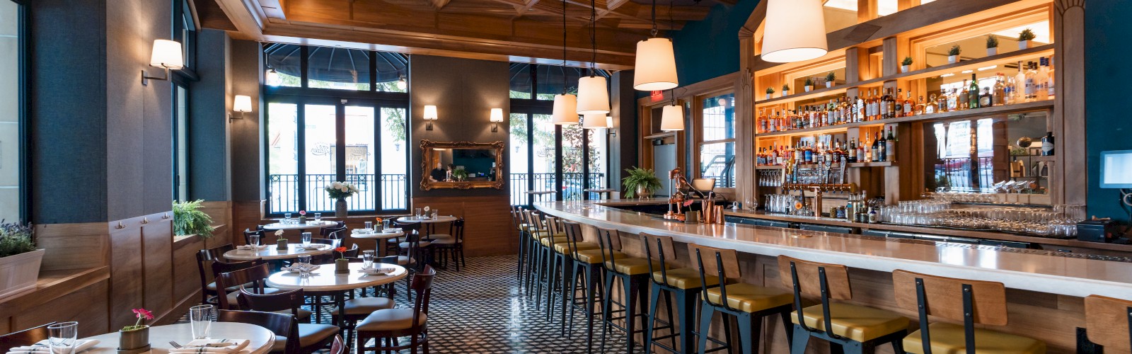 The image shows a cozy restaurant interior with a long bar, high chairs, tables, checkered floor, and pendant lights, creating a warm atmosphere.