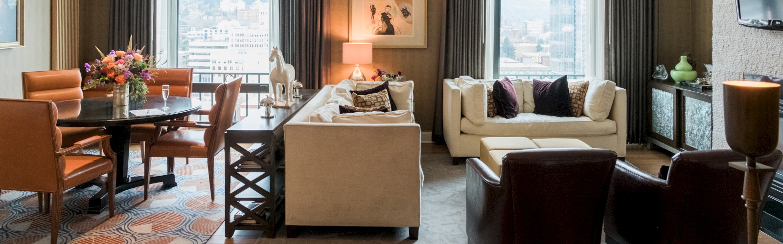 A stylish living room with a dining area, sofas, chairs, a wall-mounted TV, artwork, and large windows with city views.