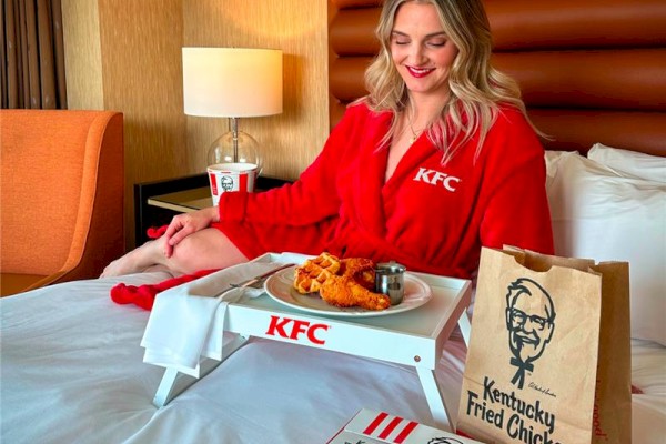 A person in a red KFC robe enjoys fried chicken and waffles on a tray in bed, with KFC packaging and a drink nearby.