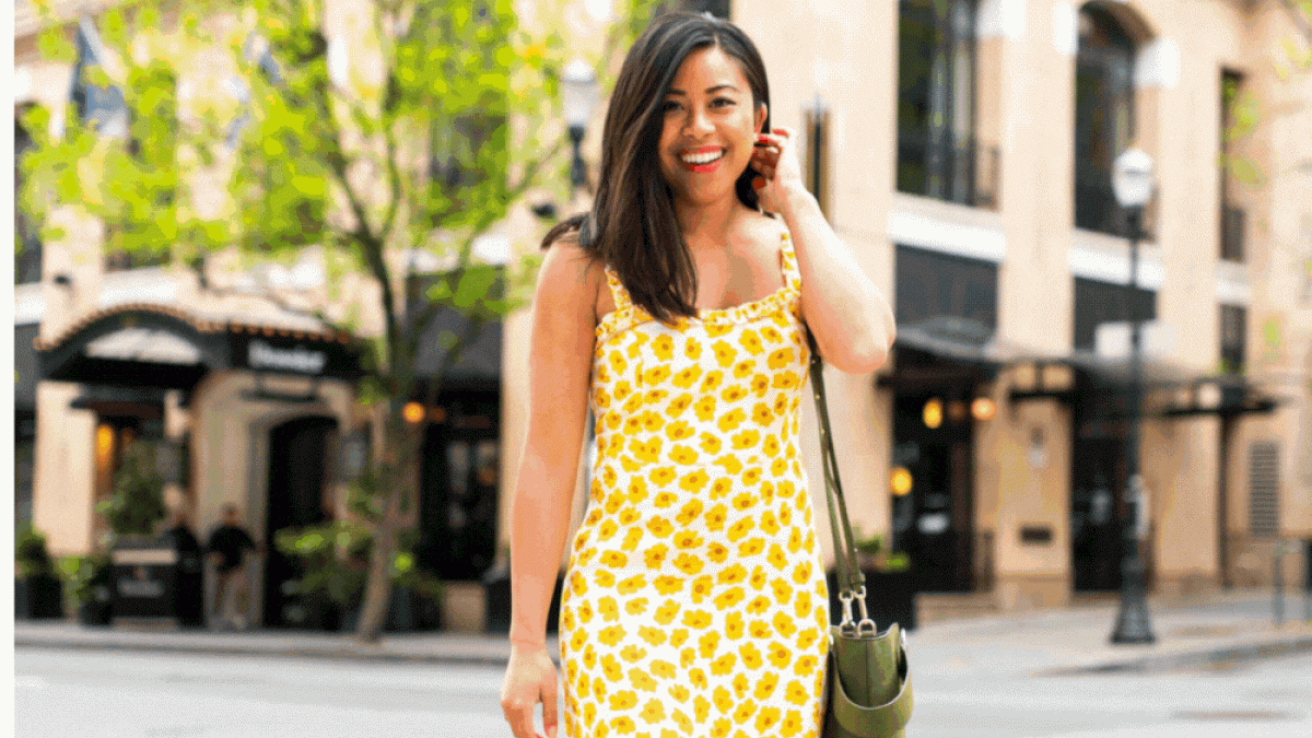 A woman in a yellow dress with a leaf pattern stands on a street, smiling and touching her hair, with shops and trees in the background.