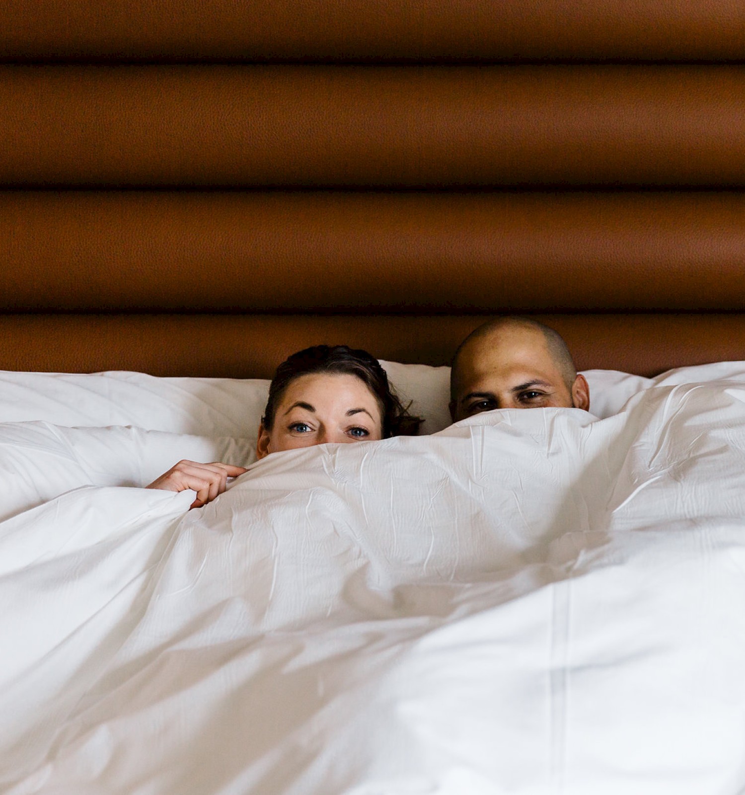 Two people are lying in bed, mostly covered by a white comforter, with only their eyes visible above the blanket.