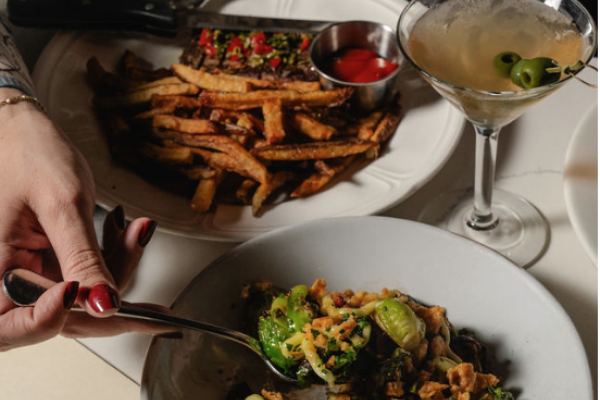 The image shows a table with a plate of pasta, a dish of fries with a dipping sauce, and a cocktail with green olives.
