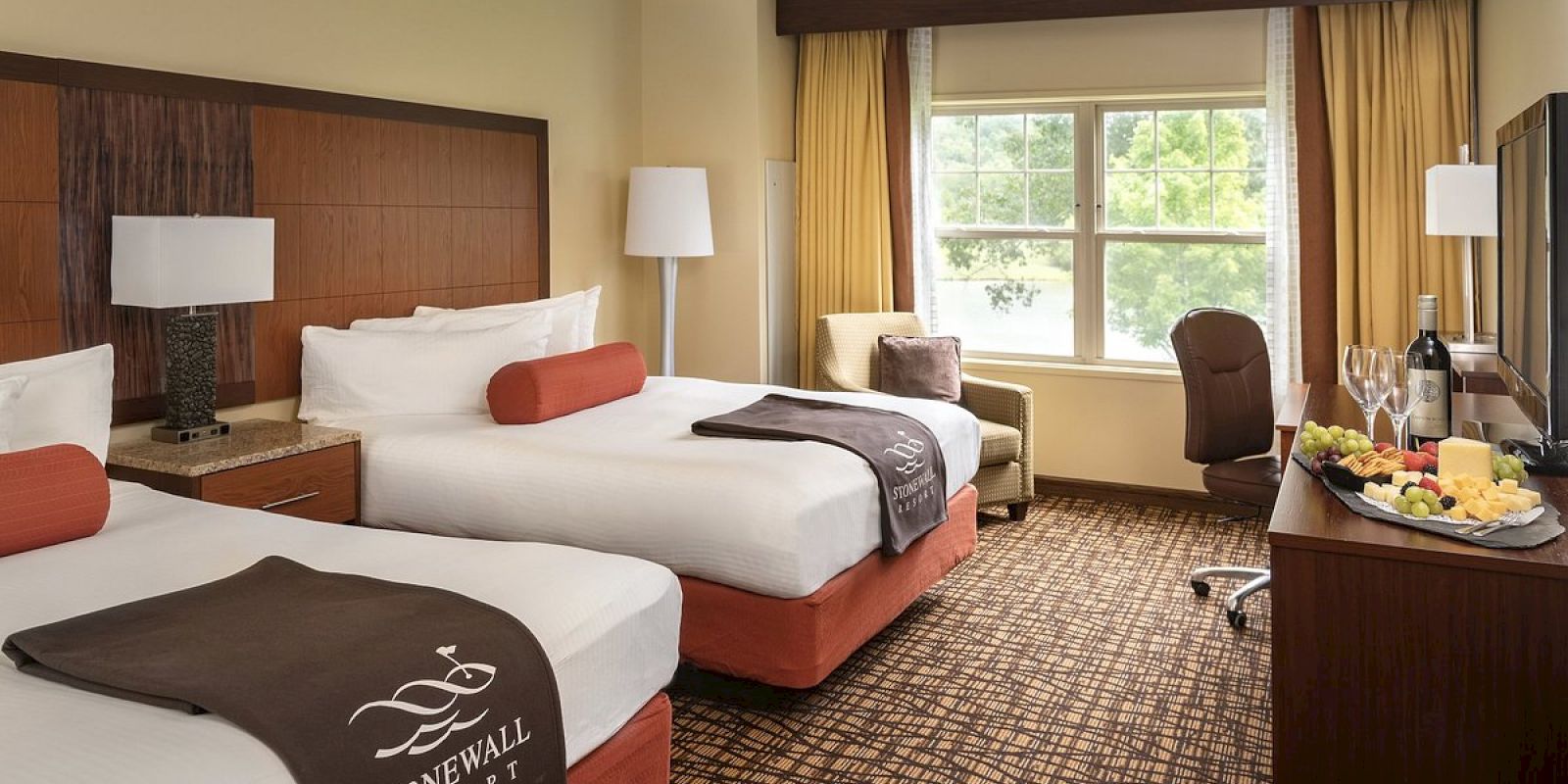 A tidy hotel room with two beds, a window, artwork, and a snack tray.