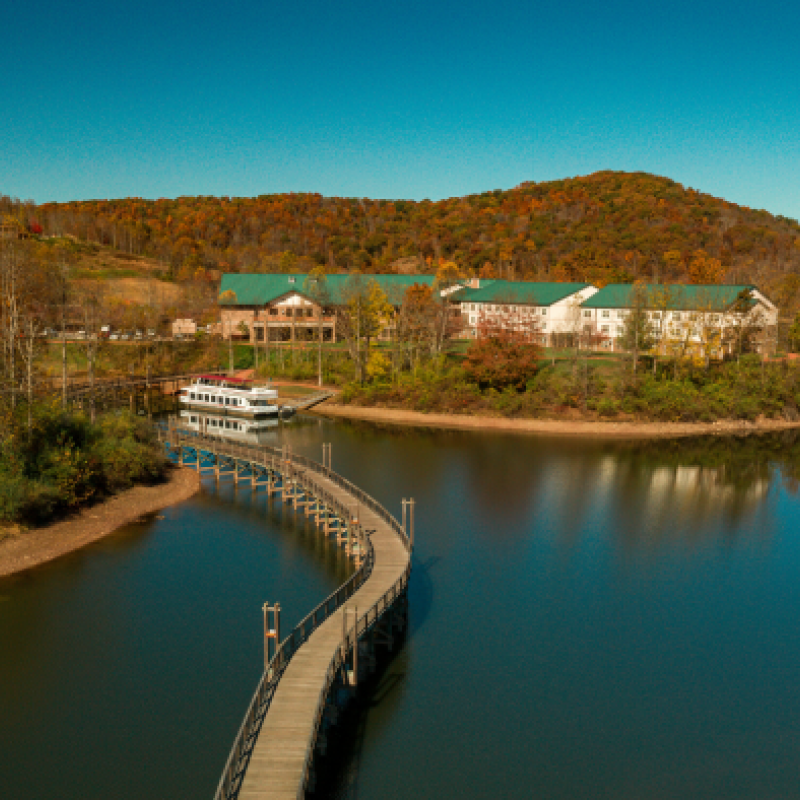 A scenic lake with a pier leading to a boat, surrounded by trees and buildings.