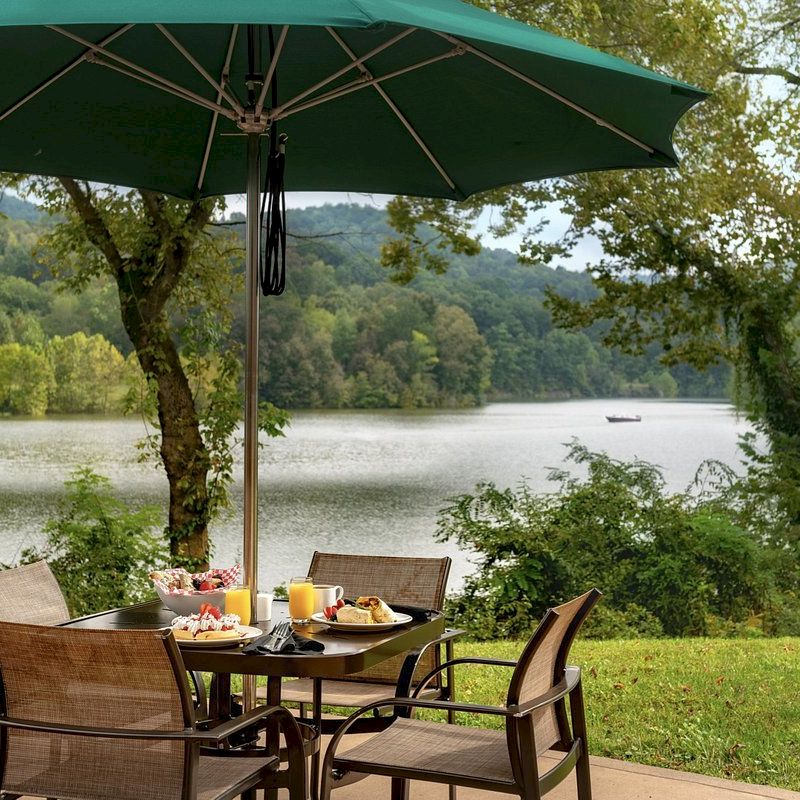 Outdoor dining setup overlooking a serene lake, with an umbrella, chairs, and a meal ready.