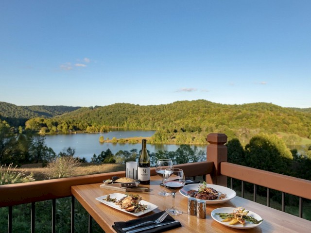 Outdoor dining setup with meal and wine; scenic lake view in the background.