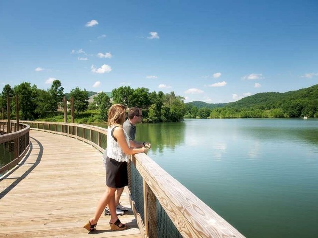 Two people stand on a lakeside boardwalk, surrounded by lush greenery and clear skies.