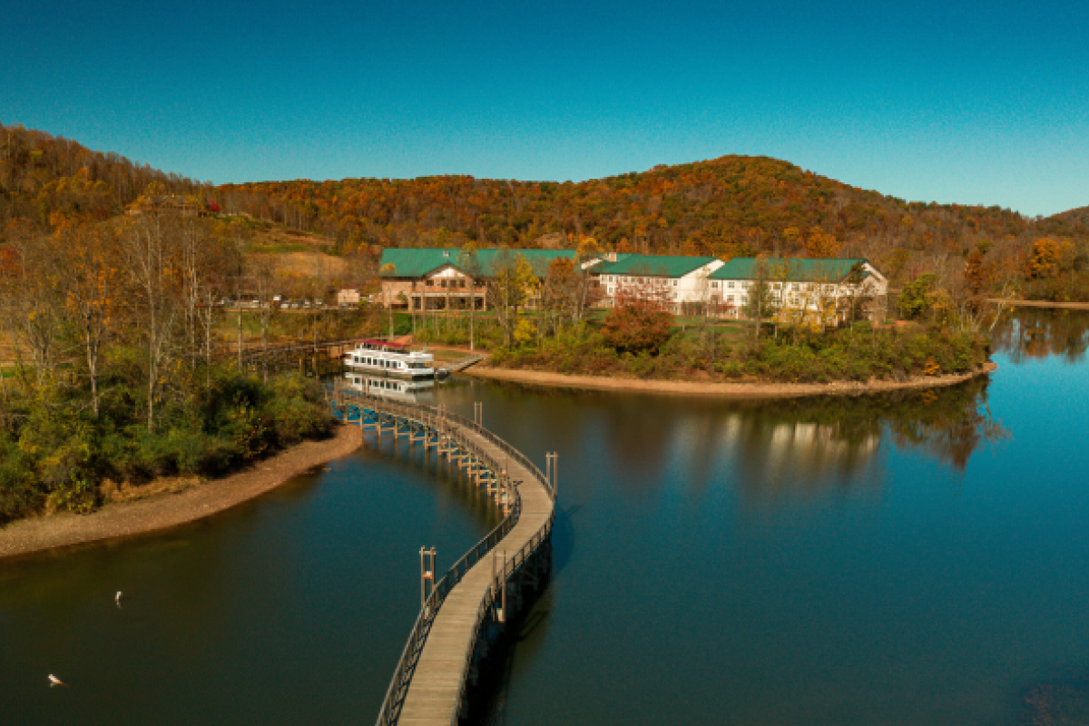 A serene lake with a long pier leading to a boat, surrounded by wooded hills and a large building.