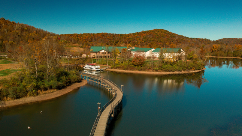 A serene lake with a long pier leading to a boat, surrounded by wooded hills and a large building.